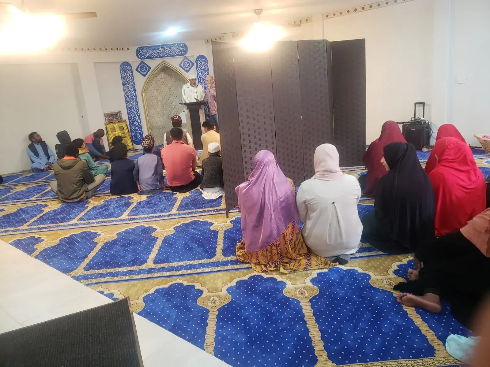 Men, women and children seated on the prayer carpet listening to a talk in the shelter's prayer hall
