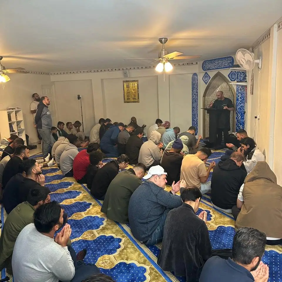 Men seated on the prayer carpet in supplication while an imam speaks in the shelter's prayer hall